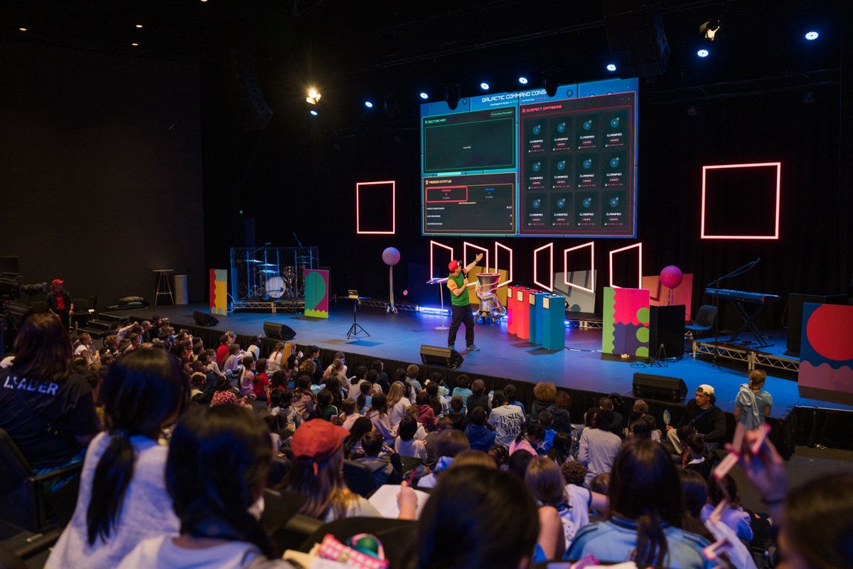 From the back of the hall: children facing a galactic-themed stage and screens.